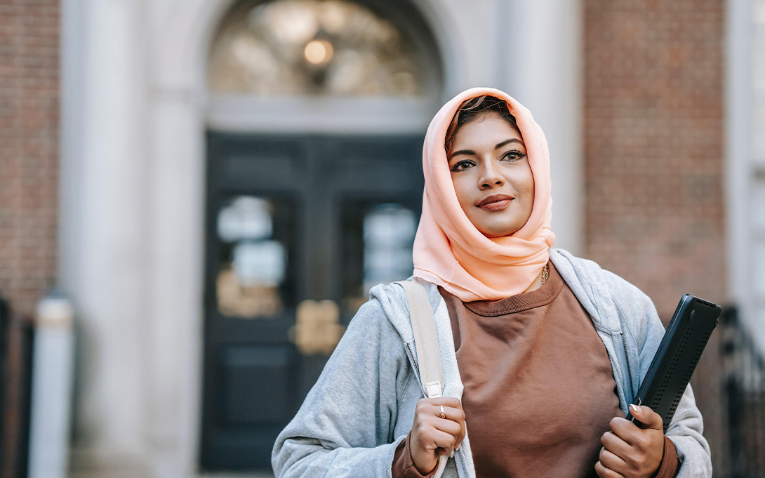 Photo of a young female student, wearing a peach-colored hijab and gray zip-up jacket, carrying a laptop and backpack