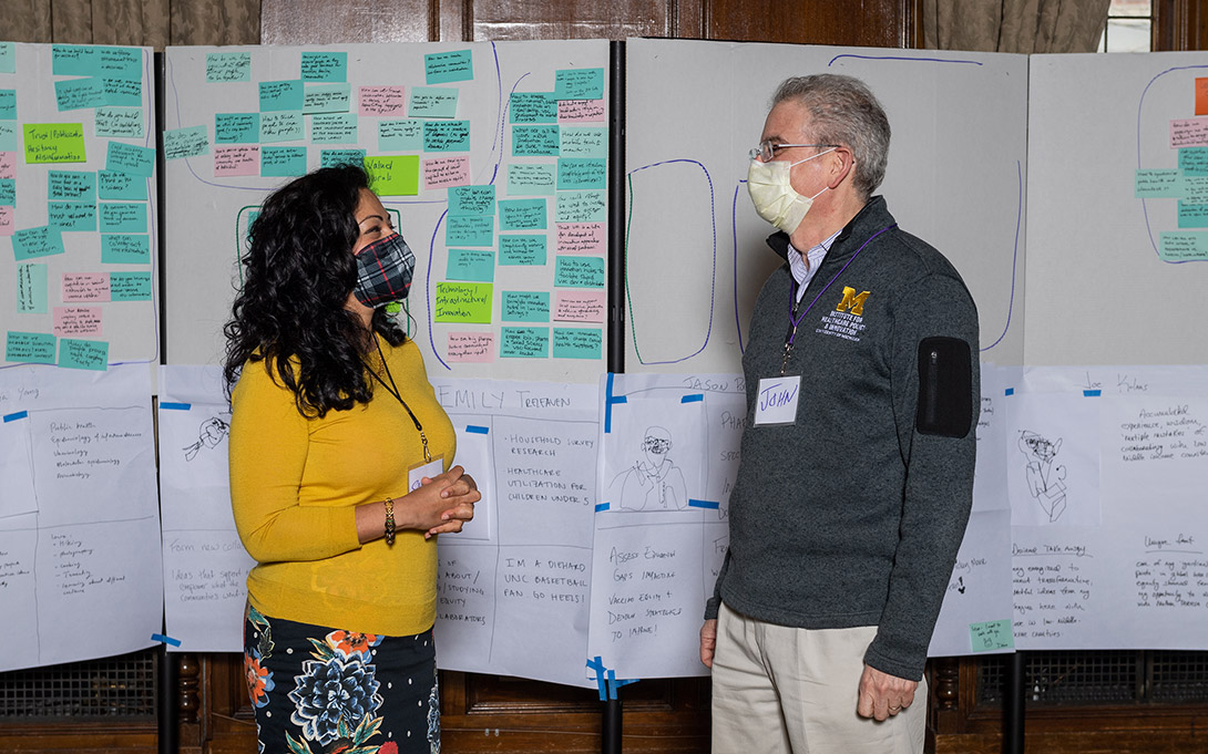 Photo of Shobita Parthasarathy facing John Ayanian, both standing in front of posters with writing and post-it notes