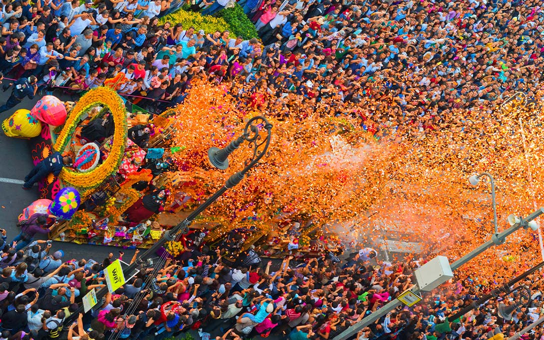 Photo, from above, of a patron saint festival in Mexico. A dense parade of people and brightly colored parade floats make their way down a local street.