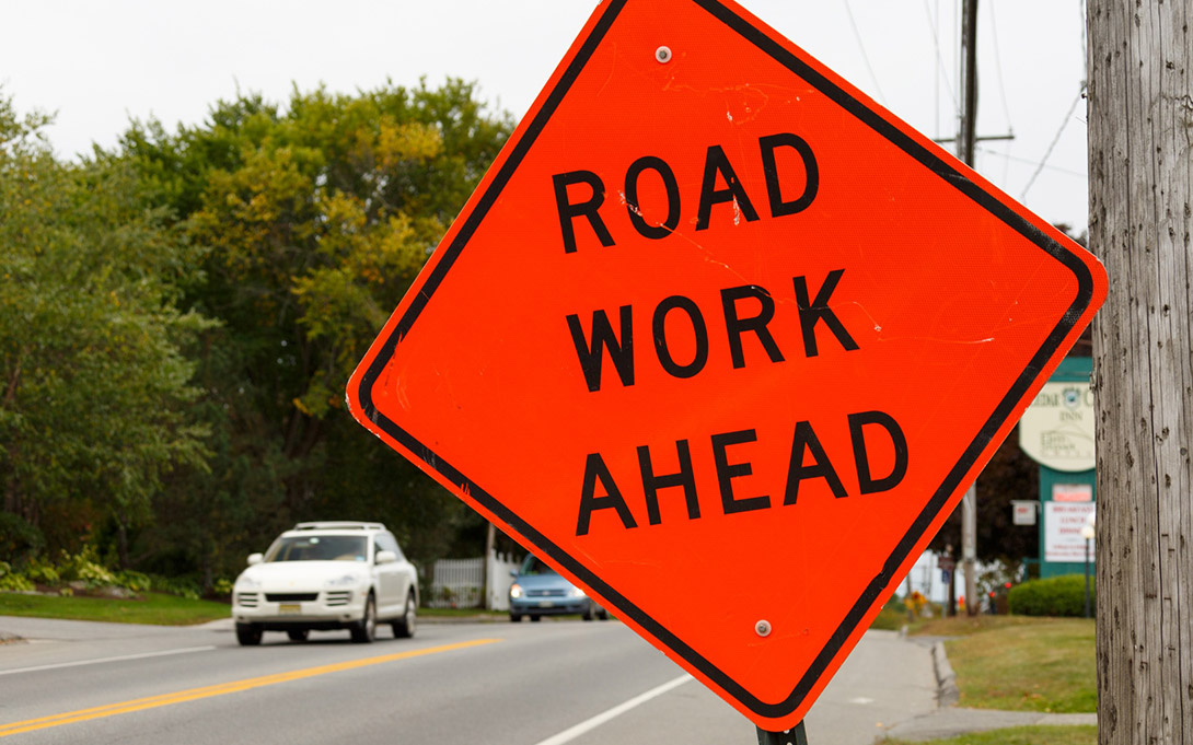 Photo of a "Road Work Ahead" sign in the foreground with cars driving along the road beside it