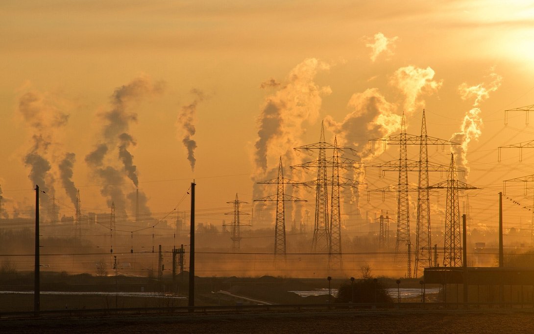 Electric towers in the near foreground dwarfed by many large plumes of smoke coming from carbon-emitting towers in the distance