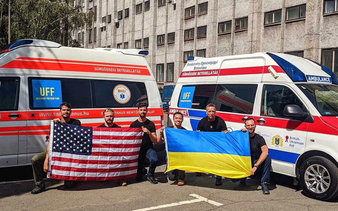 Photo of 6 men positioned in front of ambulances and holding United States and Ukraine flags