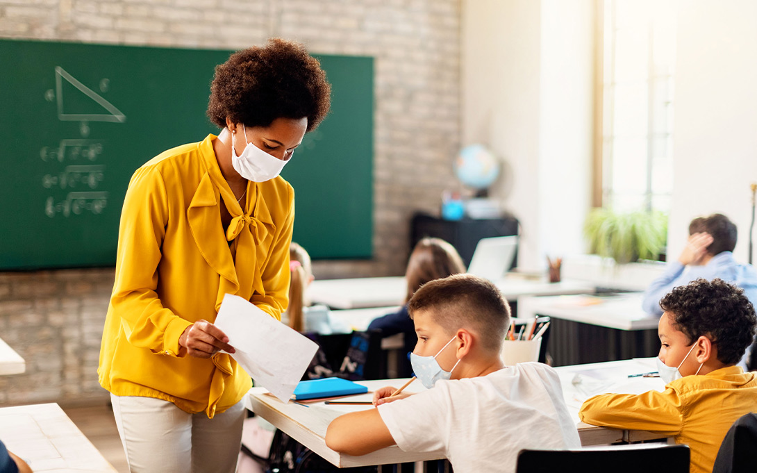 Photo of a teacher in a classroom, speaking to a young student who is seated at their desk