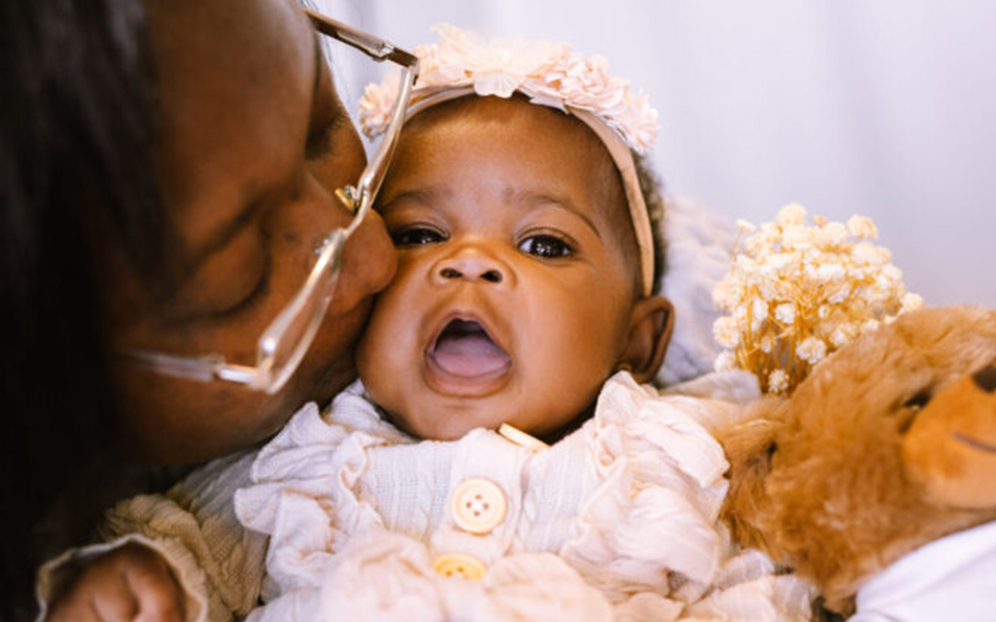 Adult kissing a baby on the cheek, the baby is holding a teddy bear and wearing a floral headband.