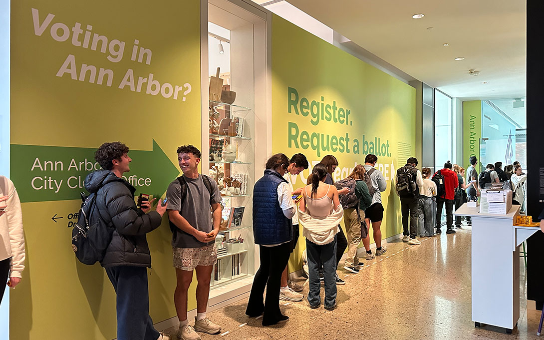 Several college-aged students line up along the hallway of the University of Michigan’s Central Campus Voting Hub. The walls are painted light green with large white and dark green text that reads, “Voting in Ann Arbor?” and “Register, Request a ballot…” alongside some other text that is obscured by the queuing students. Two students waiting in line in the foreground, look at each other talking and smiling.