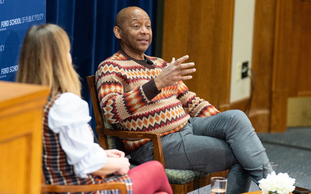 An individual wearing a colorful sweater is speaking and gesturing, seated next to another person in a discussion at the Ford School of Public Policy.