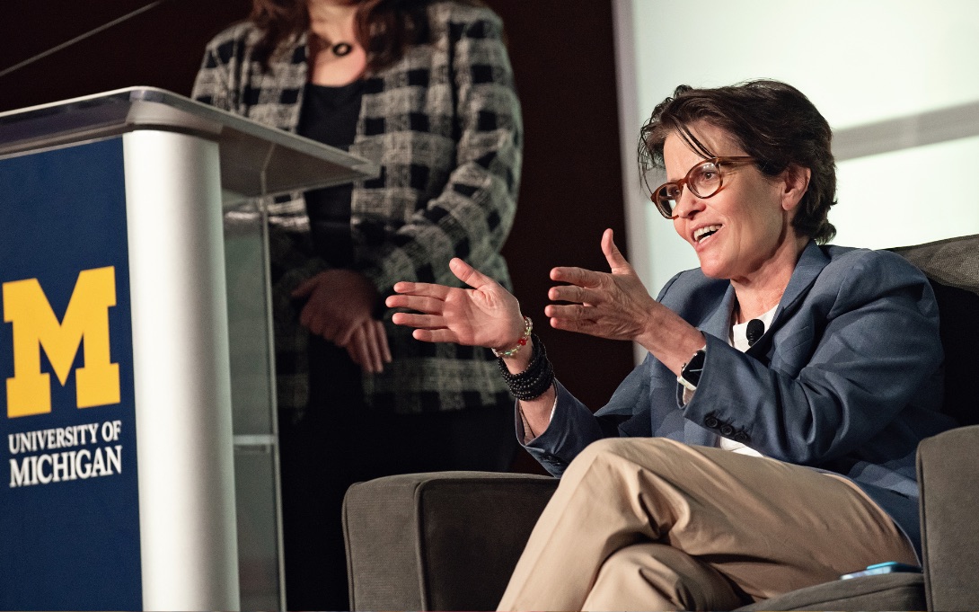 A person is speaking animatedly at a lectern with the University of Michigan logo, another individual partially visible in the background.