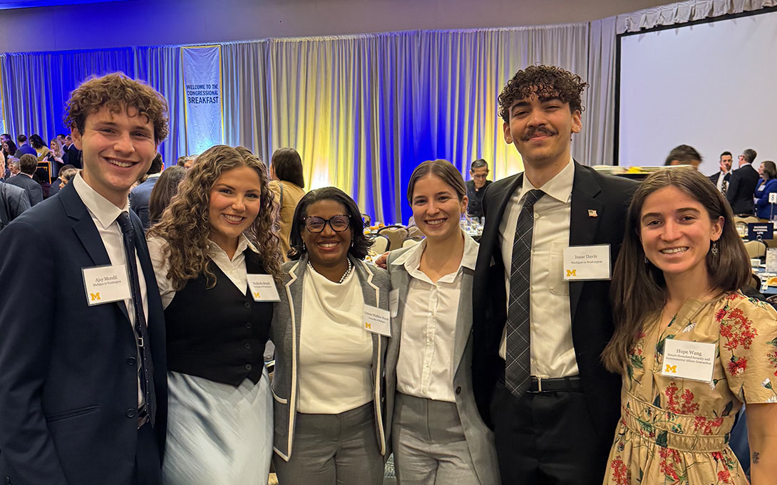 Group of six people smiling at a formal event with a banner of the Rotary International logo in the background. They are wearing name badges and formal attire.