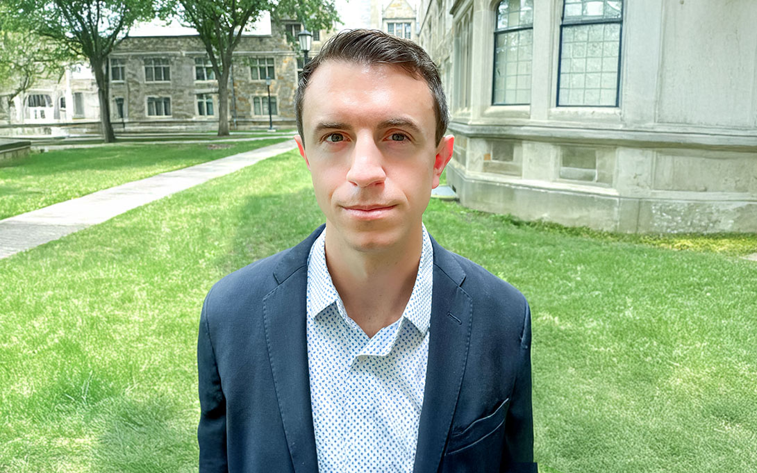 Person in a business suit sitting outside with old stone buildings and green grass in the background.