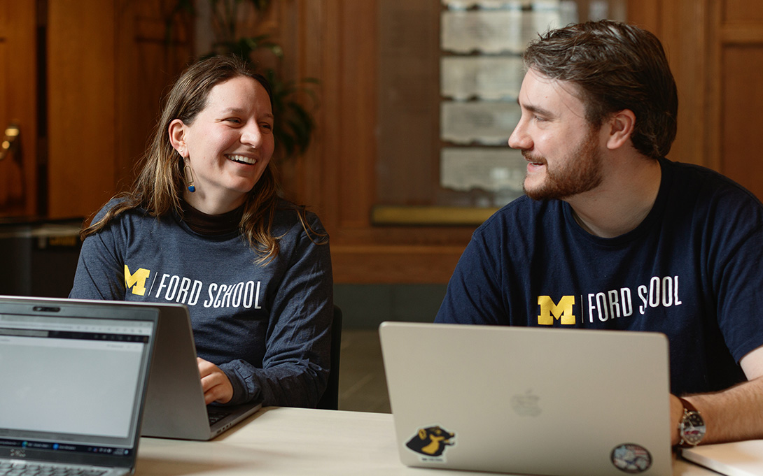 Two students wearing University of Michigan sweatshirts sit at a table, smiling and looking at each other, with a laptop open in front of them.