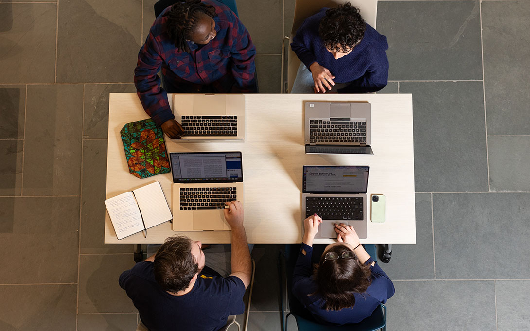 Four individuals are seated around a table, each working on a laptop. There are books and a digital tablet also visible on the table. This appears to be a collaborative work setting.