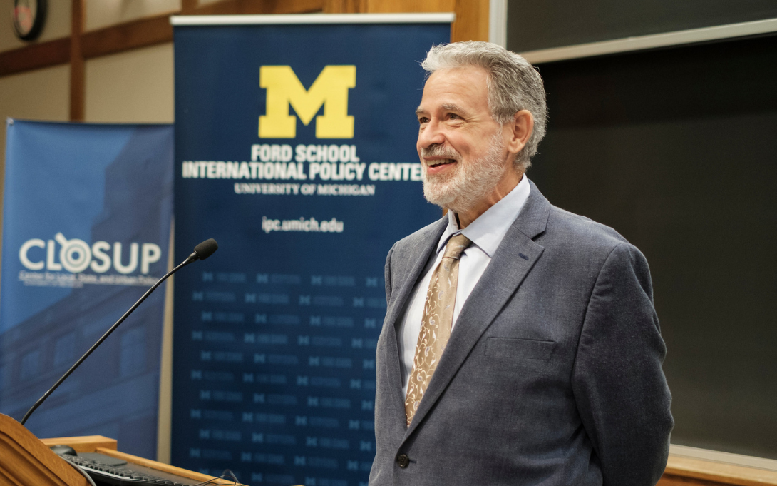 A person is smiling while standing at a podium giving a speech, with banners of the University of Michigan and Ford School International Policy Center in the background.