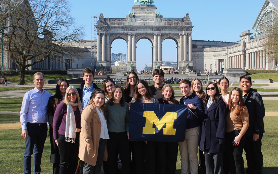 Group of people posing in front of the Cinquantenaire Arch in Brussels, holding a banner with the University of Michigan logo