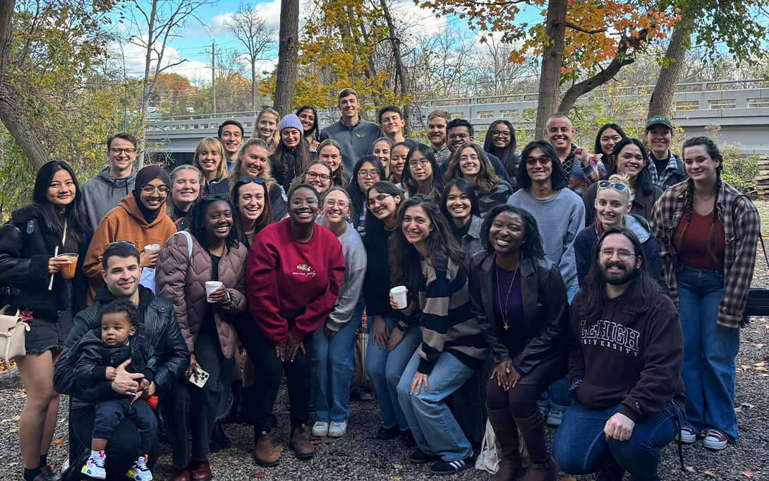 A diverse group of people smiling in an outdoor setting, with trees in the background, suggesting a casual gathering or event. Several individuals are holding beverages.