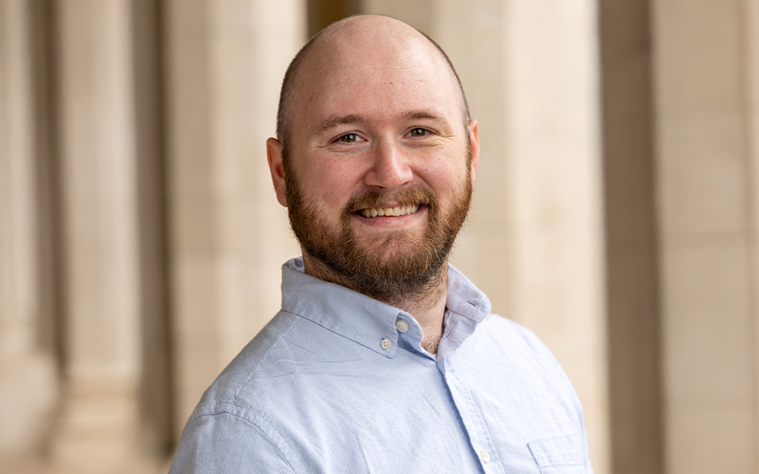 An individual smiling, dressed in a light blue shirt, with a columned architectural background.
