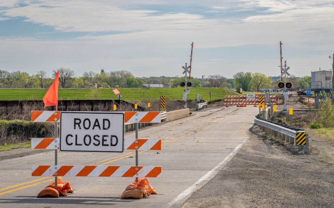 Road closed sign and barriers blocking a road under construction, with railway crossing and clear skies in the background.