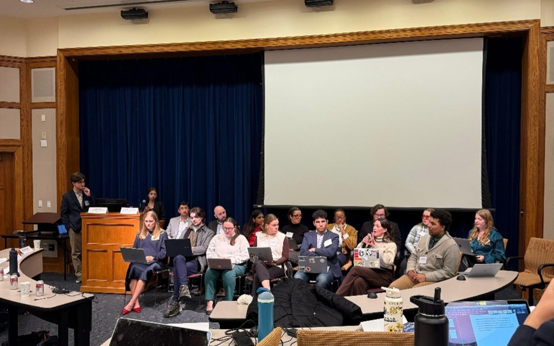 A panel discussion in progress inside a university lecture hall with multiple attendees seated and one person standing at the podium.