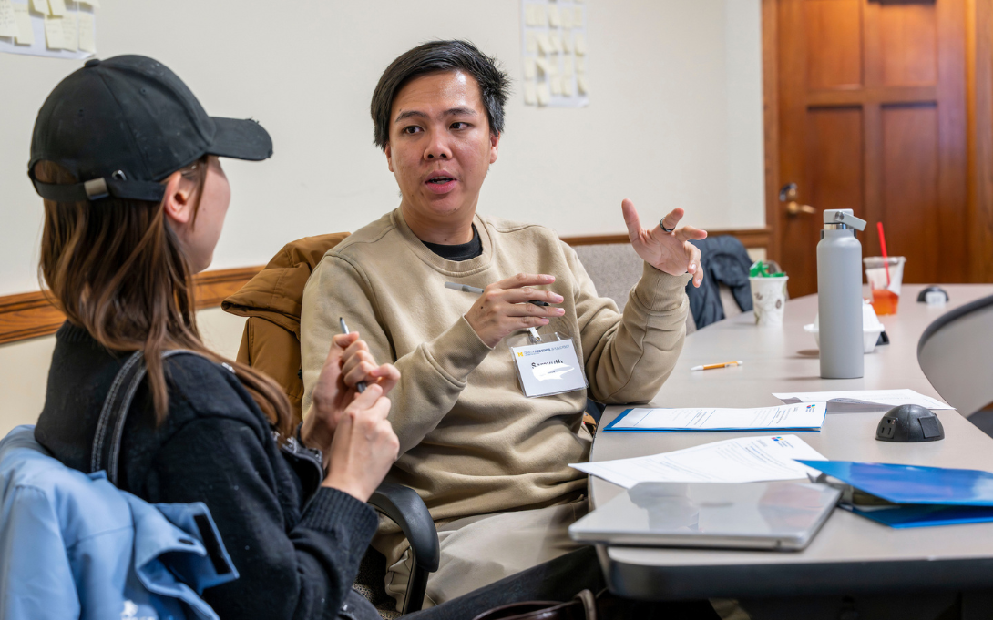 Two individuals engaged in a conversation while seated at a table with documents and a laptop. One person is gesturing with their hands while speaking to the other, who is holding a pen and listening attentively.