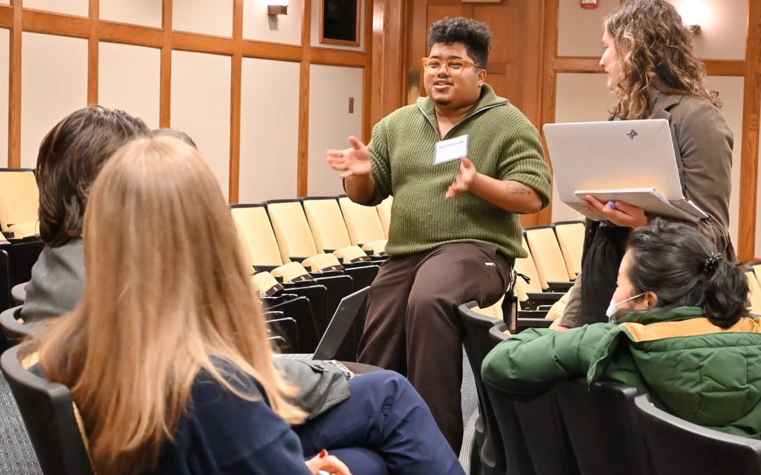 A group of people engaged in a discussion in a lecture hall. One person is standing and gesturing while speaking, another is holding a laptop and looking at the speaker. Several attendees are seated, listening attentively.