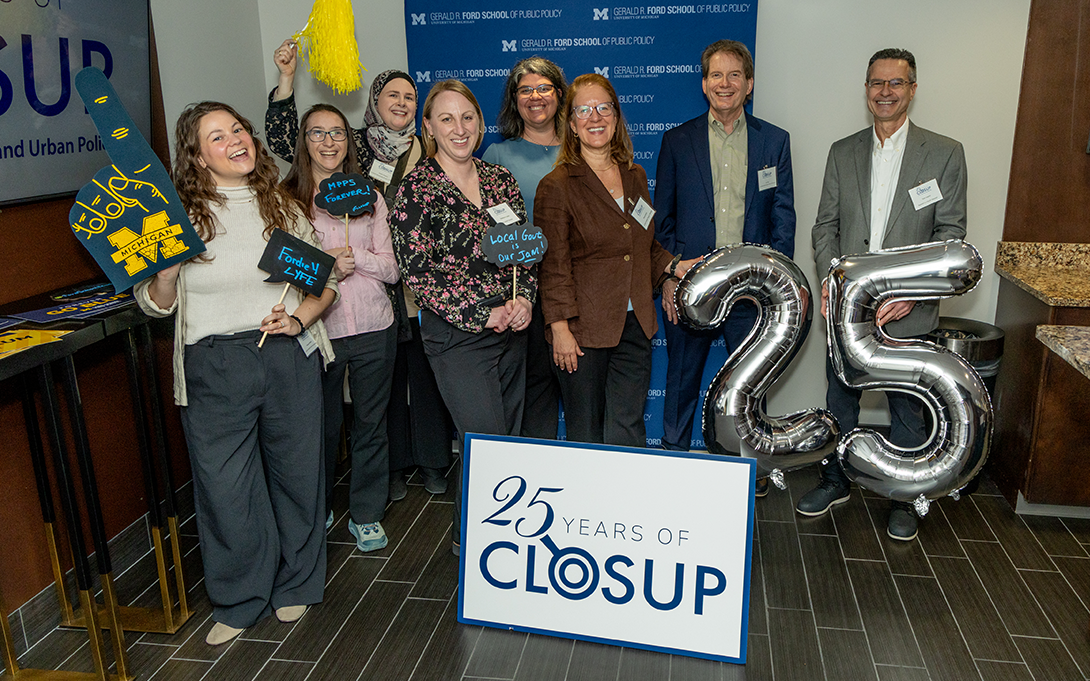 A group of people in front of large '25' balloons, holding celebratory signs, foam fingers, and pom poms