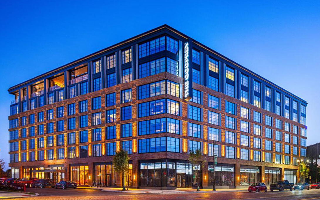 The Godfrey Hotel in Detroit, Michigan. A modern brick-and-glass hotel illuminated at dusk, shown from a street corner with cars parked along the curb. The hotel is the location of the upcoming Ford School Kohn Conference.
