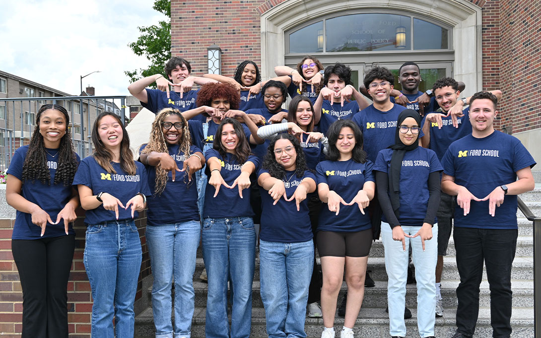 Ford School students wearing navy T-shirts pose on the steps outside the Ford School building, making block “M” hand signs.