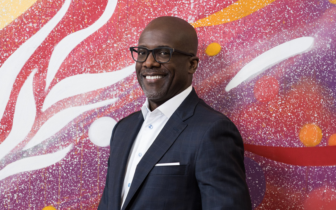 A smiling person wearing glasses and a dark suit stands before a vibrant abstract mural in red, purple, orange, and white.
