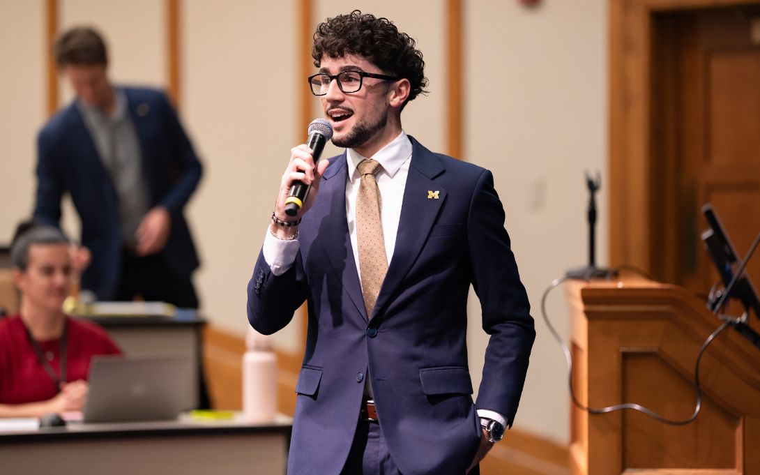 A man wearing glasses, a navy suit, and a University of Michigan lapel pin speaks into a handheld microphone in a lecture hall.
