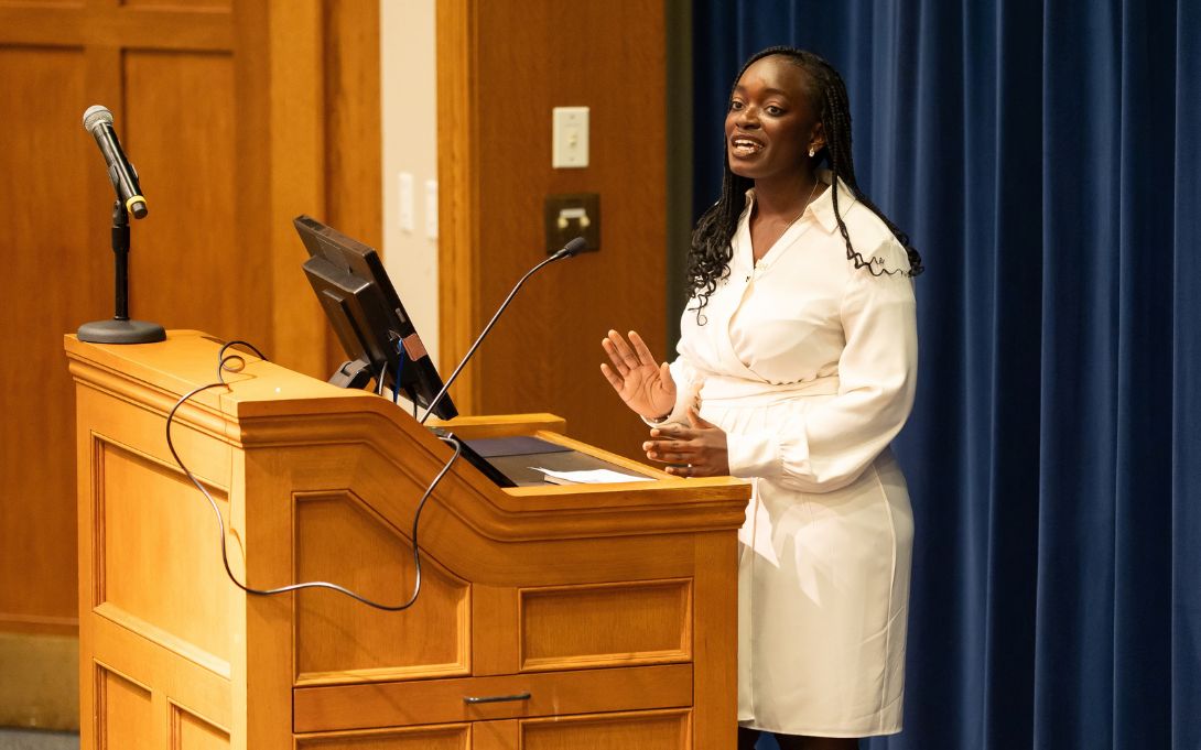A woman in a white dress speaks and gestures from behind a wooden lectern equipped with microphones and a monitor.