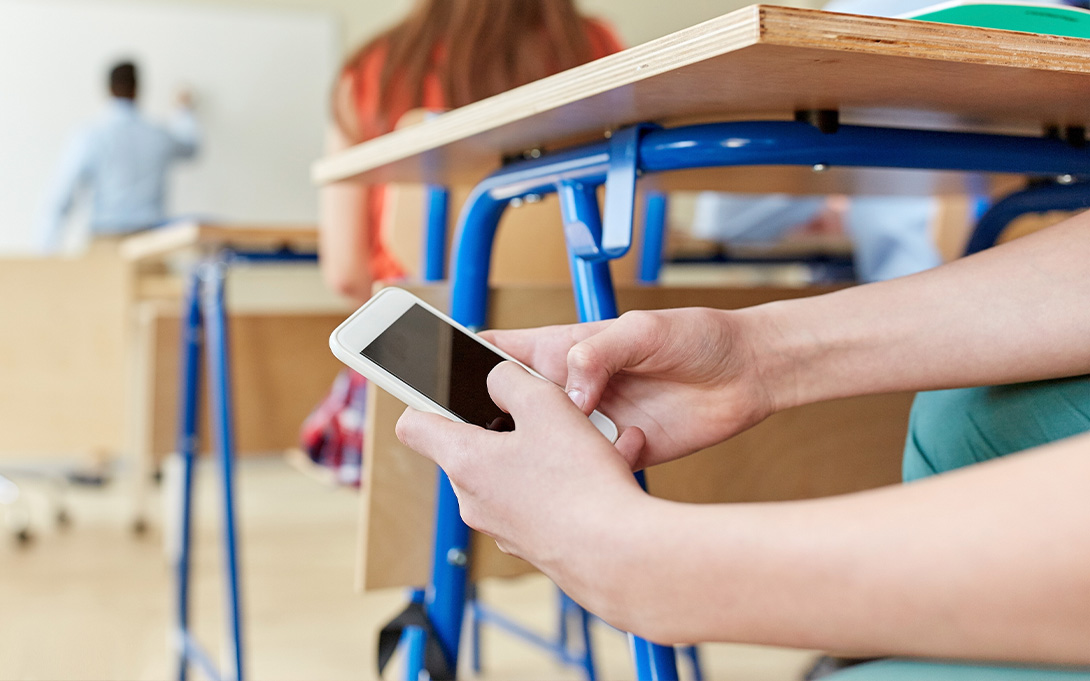 A student holds a smartphone beneath a classroom desk while other people appear blurred in the background.