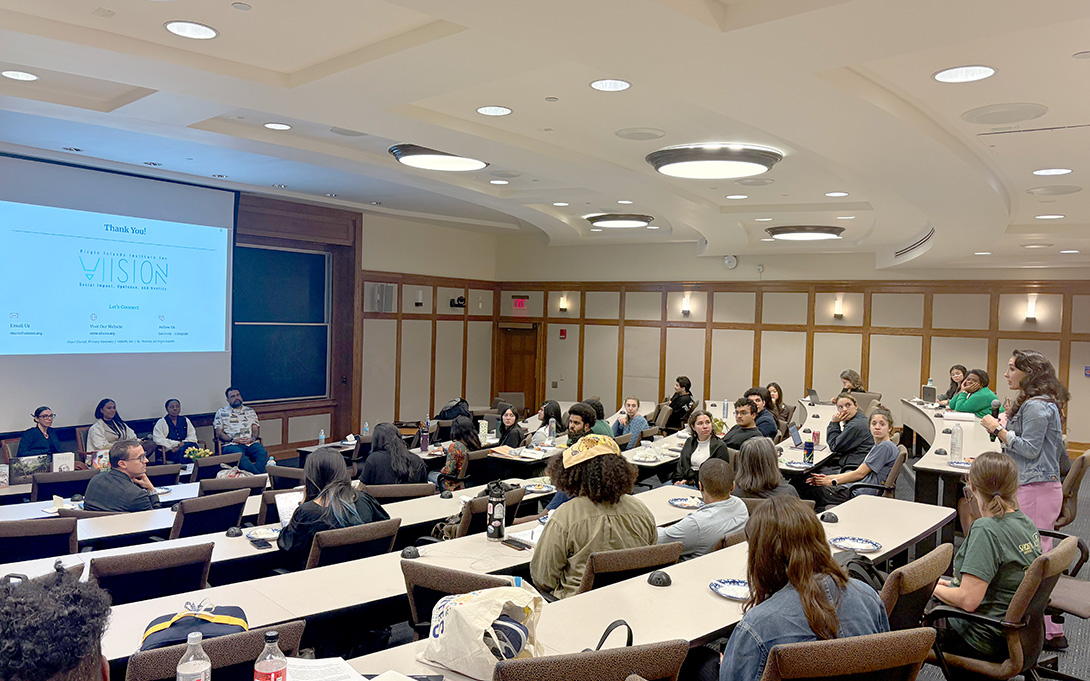 A speaker addresses attendees seated in a tiered lecture hall during a panel discussion, with a “Vision” presentation slide projected at the front.