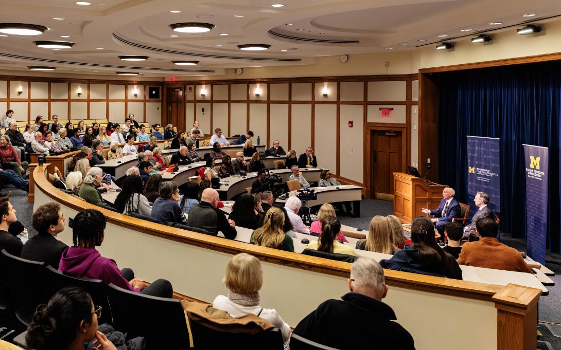 A large audience listens to two seated speakers during a University of Michigan Ford School of Public Policy event in a tiered lecture hall.