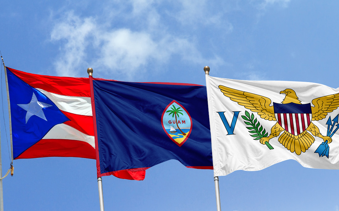 The flags of Puerto Rico, Guam, and the U.S. Virgin Islands wave on flagpoles against a bright blue, partly cloudy sky.