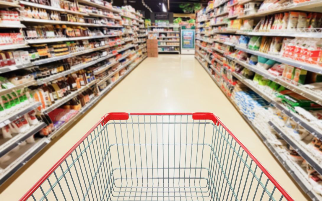 Empty shopping cart in a brightly lit grocery store aisle lined with stocked shelves.