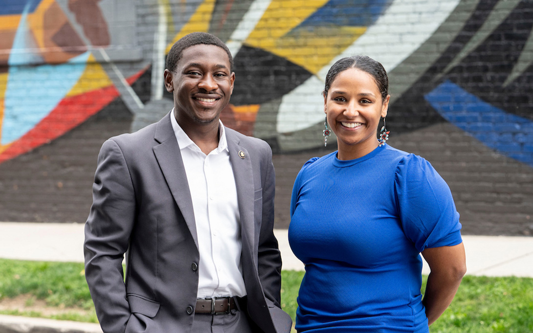 Two smiling people pose outdoors in front of a colorful abstract mural; one wears a gray suit, and the other wears a bright blue shirt.
