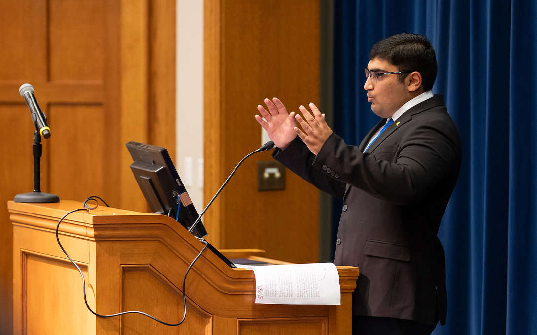 A presenter in a suit and glasses gestures while speaking at a wooden lectern beside a blue curtain.