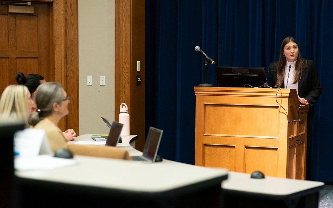 A presenter speaks from a wooden lectern as seated attendees listen in a lecture hall.