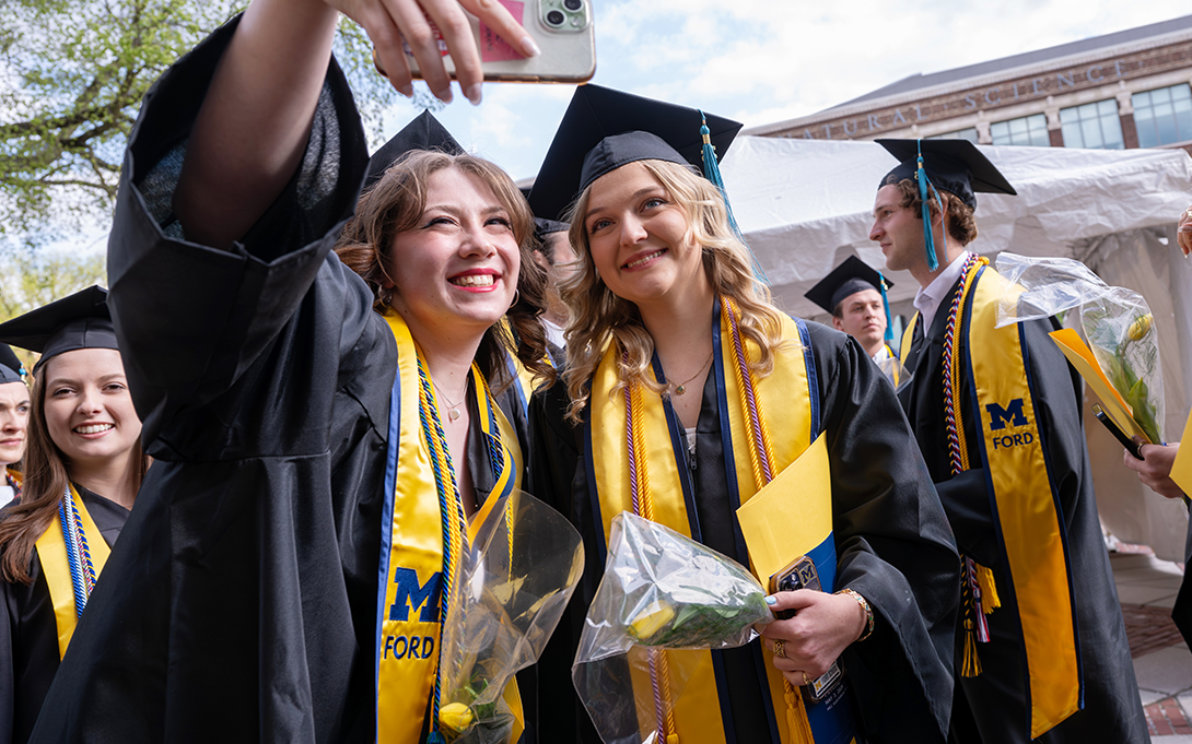 Ford School grads in caps, gowns, and maize stoles squeeze in for a selfie, yellow tulips in hand, as classmates celebrate around them.