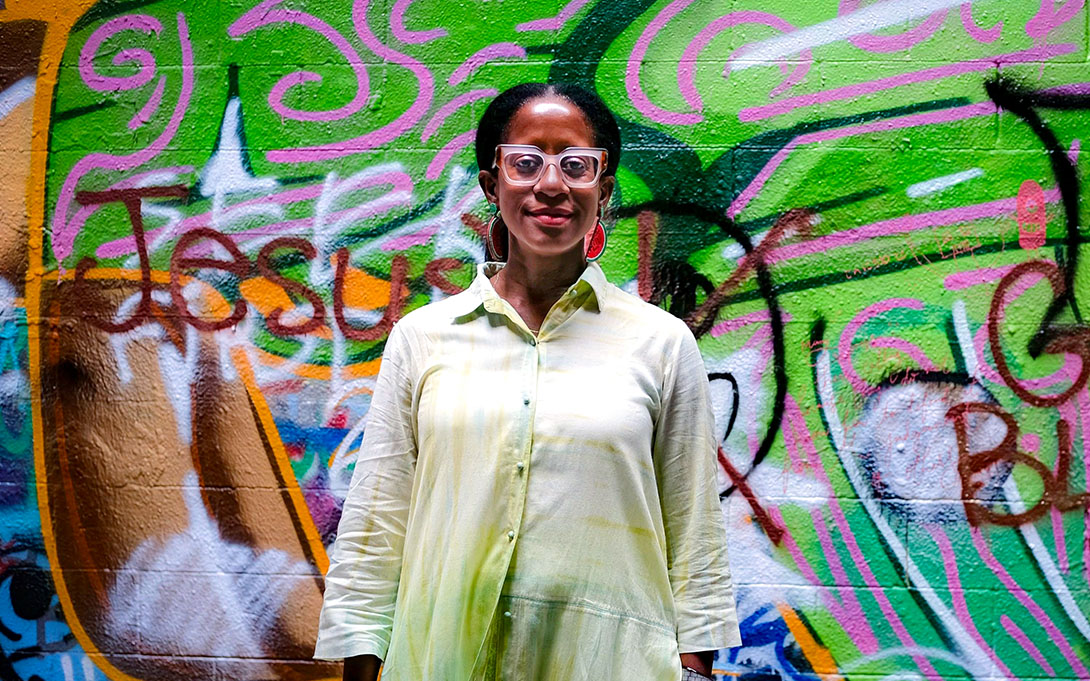 A smiling person wearing white-framed glasses and a light-colored button-up shirt stands in front of a vibrant green, pink, and multicolored graffiti wall.