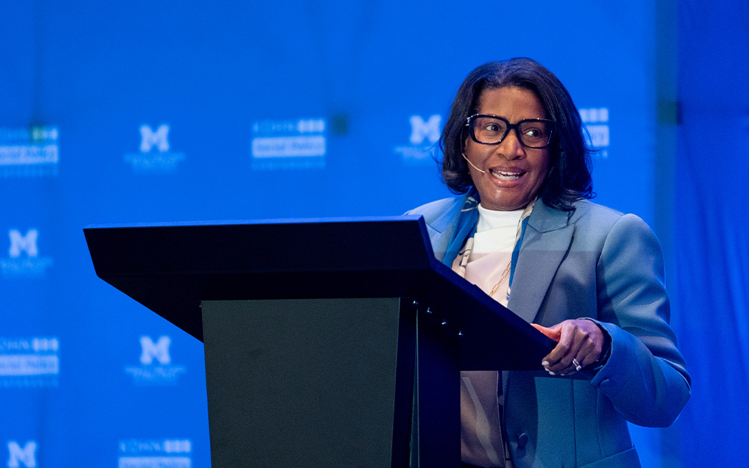 A smiling speaker wearing glasses and a blue blazer addresses an audience from behind a podium, using a headset microphone against a blue University of Michigan backdrop.