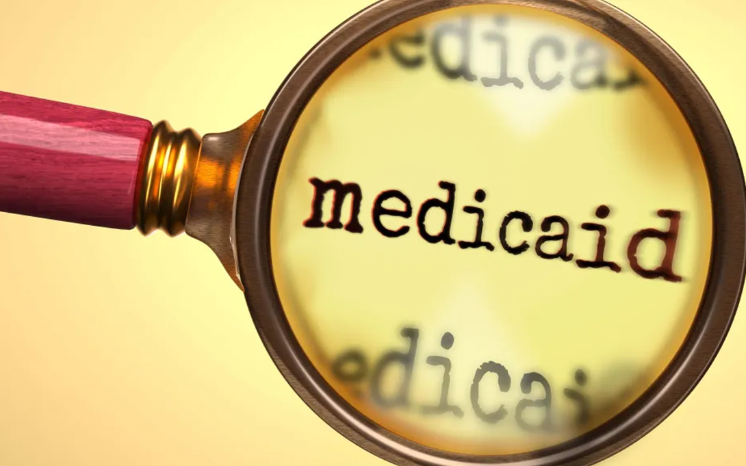 A magnifying glass enlarges the word “Medicaid” on a pale yellow background.