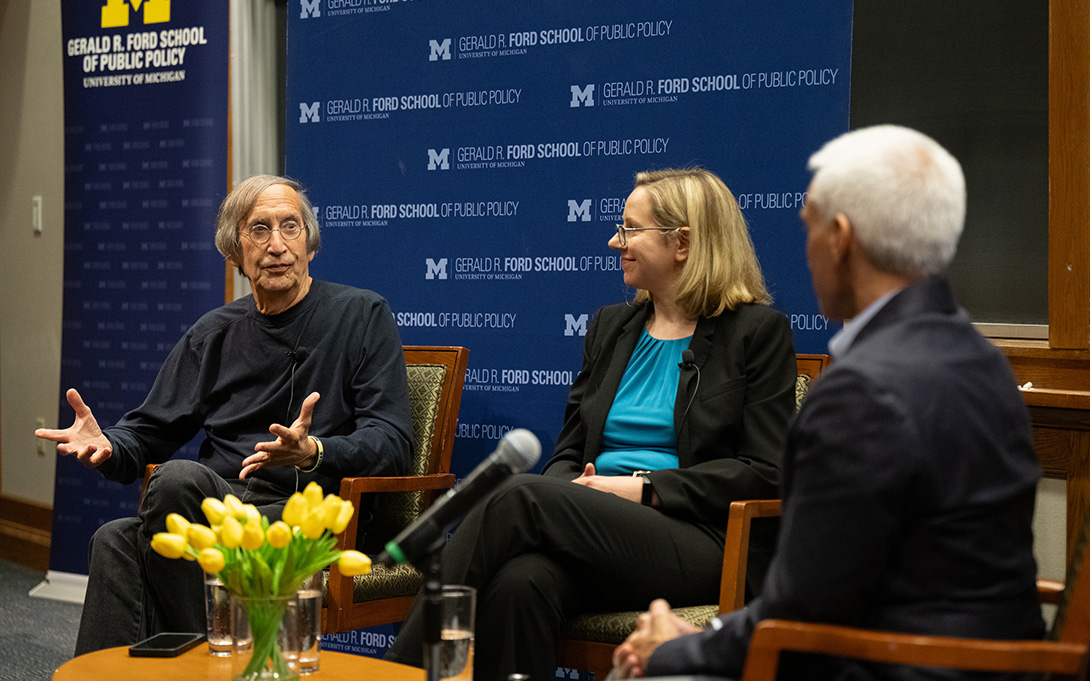 Three panelists sit onstage during a discussion at the University of Michigan’s Gerald R. Ford School of Public Policy. One panelist gestures while speaking as the others listen; yellow tulips and a microphone sit in the foreground.