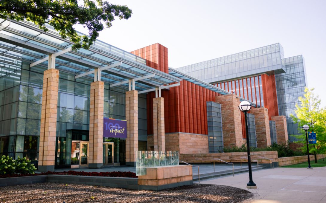 Exterior of a modern university building featuring glass walls, red and tan stone panels, a covered entrance, steps and ramps, and tree-lined walkways.