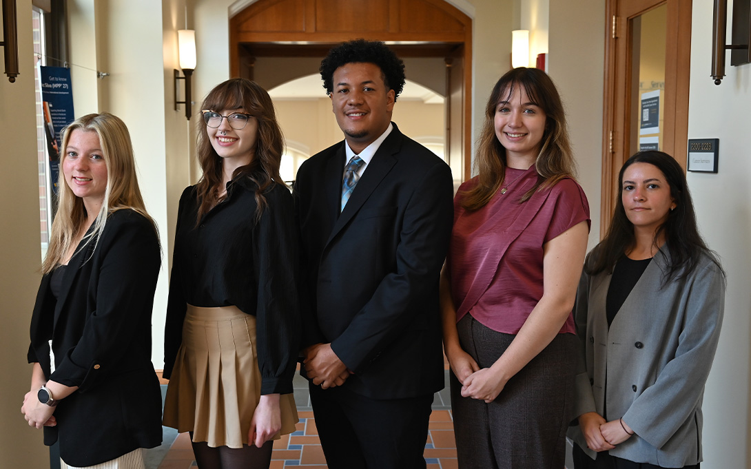 Five people in professional attire stand side by side and face the camera in a warmly lit hallway.