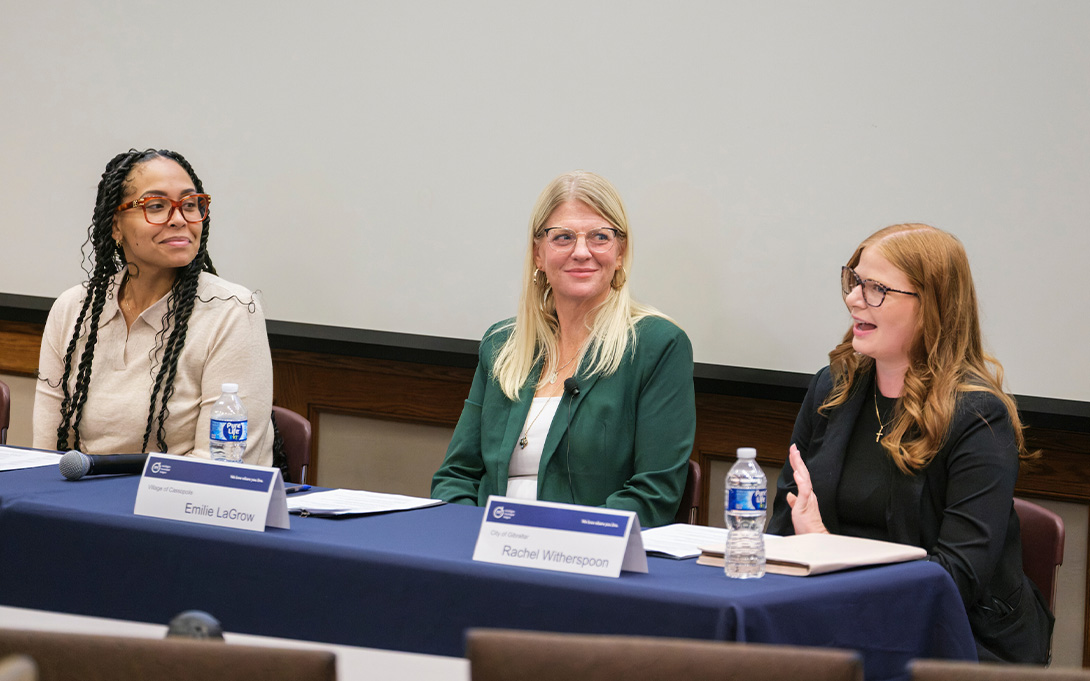Three panelists sit behind a table as one speaks and gestures while the others listen. Name placards, water bottles, notes, and a microphone are arranged on the table.