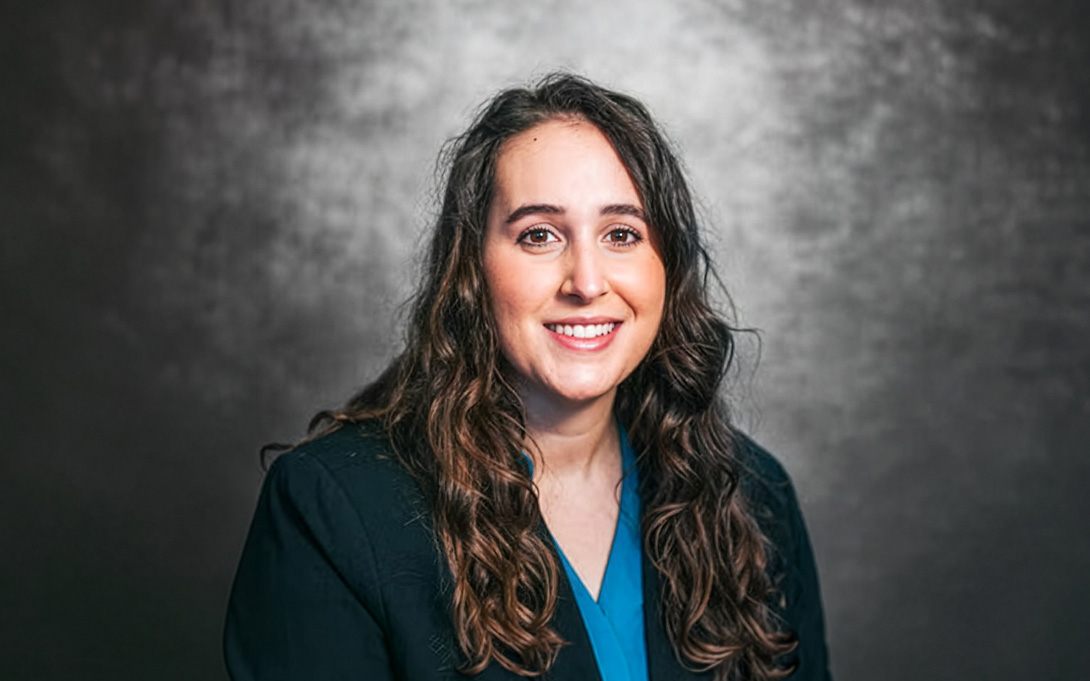 A smiling person with long, wavy brown hair wears a dark blazer and blue shirt against a textured gray background.