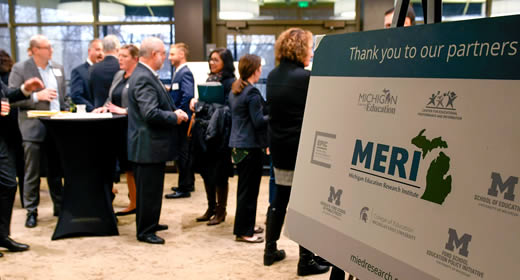 Photo of a posterboard of MERI partners in the foreground; with kickoff event attendees networking in the background
