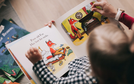 Photo of a young child, held in the lap of an a guardian or teacher, reading a book