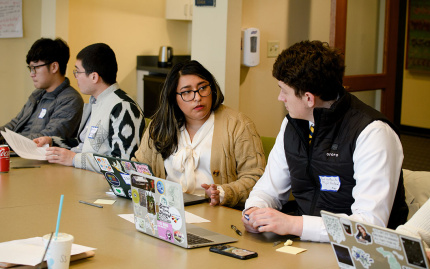 Four people sit at a long table, the two on the right with their laptops open and engaging in a discussion. The two on the left look off camera to the left