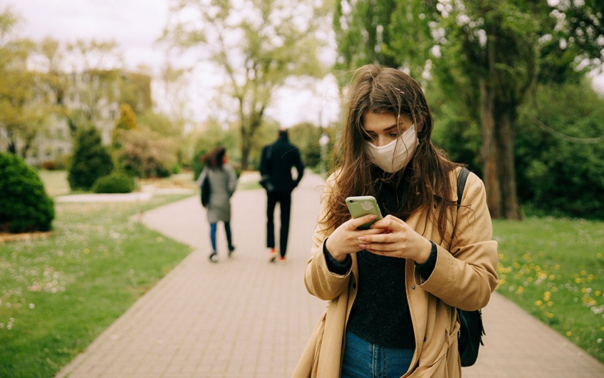 A young woman wearing a face mask stops on the sidewalk to look at her phone